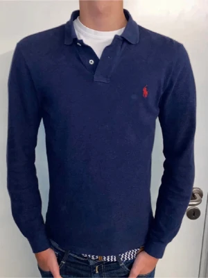 Mörkblå långärmad piké från Ralph Lauren - Mörkblå långärmad pikétröja från Ralph Lauren med klassisk krage och knappar vid halsen. Modellen är 190 och väger 73 kg, men tröjan passar som bäst om man är runt 175-185. Tröjan är använd sparsamt, skick = 8/10. Inga deffekter finns