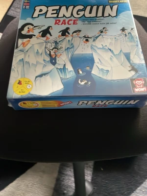 Penguin Race sällskapsspel - Roligt sällskapsspel för barn och familj där pingviner tävlar på isberg. Lådan är blå med illustrationer av pingviner och is. Perfekt för spelkvällar och passar 2-4 spelare. Speltid ca 15-30 minuter.