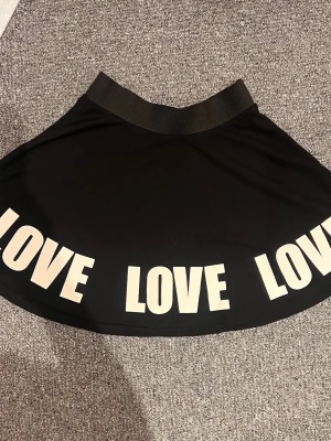 Svart kortkjol med LOVE-tryck - Cool svart kortkjol med bred resår i midjan och stora vita bokstäver där det står 'LOVE' runt hela nederkanten. Kjolen har en lätt utställd form och är perfekt för dig som gillar statement-plagg.