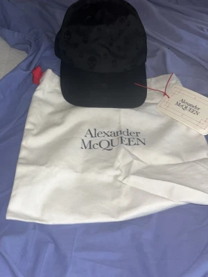 Svart keps från Alexander McQueen - Snygg svart keps från Alexander McQueen med diskret dödskallemönster över hela kepsen. Klassisk böjd skärm och justerbar passform. Tillverkad i ett glansigt material som ger en exklusiv känsla. Perfekt för dig som gillar streetstyle och designeraccessoarer.