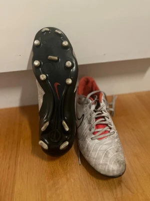 Nike vita och röda fotbollsskor - Säljer ett par vita fotbollsskor från Nike med röda detaljer och svart swoosh. Skorna har klassisk snörning, rödfärgat innerfoder och dobbar i plast för bra grepp på planen. Perfekta för dig som vill ha stilrena och funktionella skor till match eller träning.
