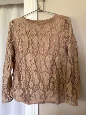Beige spetstop med blommönster - Beige top med långärmade spetsdetaljer och blommigt mönster. Framsidan är helt i spets med ett tunt foder under, medan baksidan är slät. Rund halsringning och normal passform. Perfekt för en romantisk och trendig look.