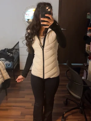 Vit väst - Snygg vit dunväst med svart dragkedja och detaljer. Västen har en figurnära passform och quiltad design som ger en modern look. Perfekt att slänga över en hoodie eller långärmad tröja för extra värme. Materialet är lätt och smidigt.