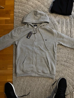 Grå zip hoodie från Polo Ralph Lauren - Snygg grå zip hoodie från Polo Ralph Lauren med liten broderad logga på bröstet. Klassisk modell med huva och dragsko, två fickor framtill och ribbade muddar. Tillverkad i mjuk bomullsblandning för skön känsla.