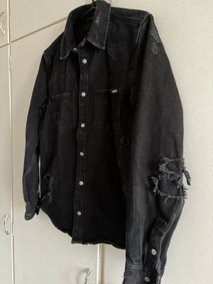 Svart distressed jeansjacka  - Säljer en svart jeansjacka  med slitna detaljer och råa kanter på ärmar och bröst. Jackan har två bröstfickor med lock, silverfärgade knappar och en avslappnad passform. Använd en gång