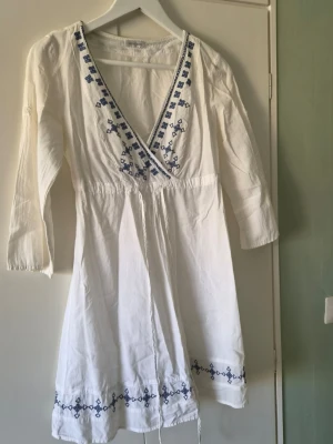 Vit omlottklänning med blå broderi - Vit omlottklänning med blå broderade detaljer längs v-ringningen och nederkanten. Klänningen har trekvartsärm och knyt i midjan för en snygg passform. Perfekt för sommarens alla tillfällen.