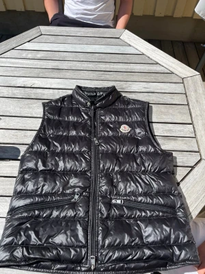 Svart dunväst från Moncler - Snygg svart dunväst från Moncler med glansig finish och klassisk logotyp på bröstet. Västen har hög krage, två dragkedjeförsedda fickor och stängs med dragkedja framtill. Perfekt för lager-på-lager och streetwear-stil.