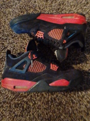Svarta och röda Air Jordan 4 sneakers - Säljer ett par Air Jordan 4 sneakers i svart mocka med röda detaljer och meshpaneler. Skorna har klassisk Jumpman-logga på plösen och svarta skosnören. Snygg retrostil med kontrasterande färger och ikoniska plastdetaljer vid snörningen.