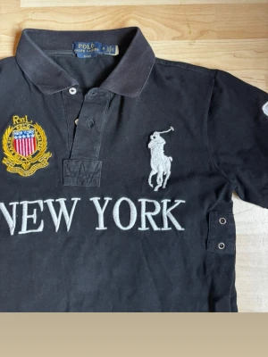Svart pikéskjorta Polo Ralph Lauren New York  - Svart pikéskjorta från Polo Ralph Lauren med stora broderade detaljer, bland annat New York-text, emblem och den klassiska polospelaren. Skjortan har korta ärmar, krage och knappar framtill. Tillverkad i bomull för en bekväm känsla. Är storlek M men passar mer som en S. Är en ganska slim modell 