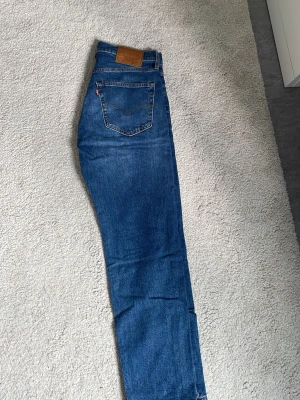 Blå raka jeans från Levi's - Klassiska blå jeans från Levi's med rak passform och femficksdesign. Jeansen har en brun läderpatch bak i midjan och tydliga sömmar. Tillverkade i slitstark denim med en snygg tvättad look. Knappt använda. 