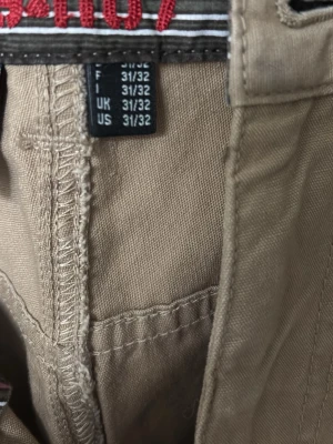 Beige chinos från Hilfiger Denim - Säljer ett par klassiska beige chinos från Hilfiger Denim. Byxorna har rak passform, fem fickor och snygga detaljer i midjan med randigt innerband och röd brodyr. Tillverkade i mjukt bomullstyg som är sköna att bära hela dagen.