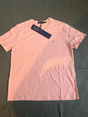 Ljusrosa t-shirt från Polo Ralph Lauren - Säljer en ljusrosa t-shirt från Polo Ralph Lauren med klassisk passform och korta ärmar. T-shirten har en liten broderad logga i ljusblått på bröstet och är gjord i mjuk bomull. Perfekt till jeans eller shorts för en clean och enkel stil.