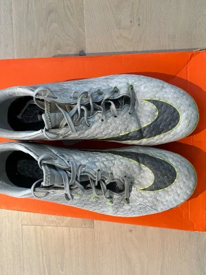 Nike grå fotbollsskor med neon - Säljer ett par grå Nike fotbollsskor med svart swoosh och detaljer i neonlime. Skorna har snörning, låg profil och dobbar för spel på gräs. Ovandelen är i syntetmaterial som ger bra bollkontroll. Perfekta för dig som vill sticka ut på planen.