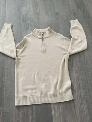 Beige stickad tröja med halv dragkedja - Snygg beige stickad tröja med halv dragkedja i halsen och ribbade muddar vid ärmslut och nederkant. Tröjan har en rak passform och hög krage som ger en clean och stilren look. Perfekt att matcha med jeans eller kjol.