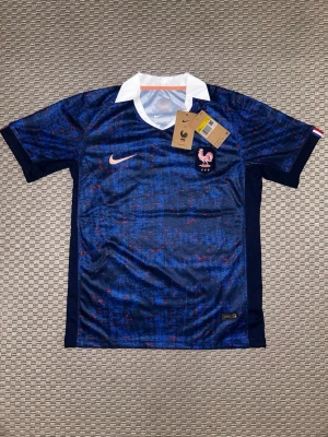 Frankrike fotball jersey - Snygg blå fotbollströja från Nike med Frankrikes landslagsemblem och vit krage. Tröjan har ett unikt mönster i blått och rött samt Nike-logga i vitt. Tillverkad i lätt och ventilerande material, perfekt för träning eller match.