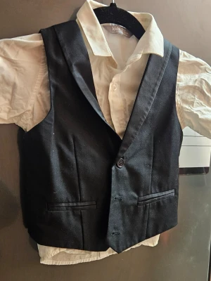 Svart väst o vit skjorta  från Lindex Kids, stl 92 - Klassisk svart väst från Lindex Kids i storlek 92. Västen har två fickor framtill, knäppning med två knappar och en stilren, tidlös look o  en vit skjorta för en dressad outfit. Materialet känns mjukt och bekvämt.