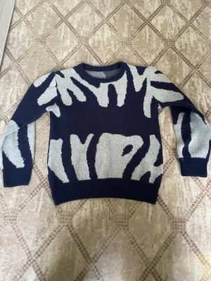Mörkblå stickad tröja med mönster - Cool mörkblå stickad tröja med abstrakt ljusgrått mönster över hela plagget. Tröjan har rund halsringning, långa ärmar och ribbade muddar vid ärmslut och nederkant. Perfekt för dig som gillar unika prints och vill sticka ut.