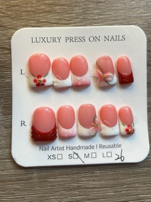 Press-on naglar rosa med blommor - Snygga press-on naglar i rosa och vitt med röda detaljer och 3D-blommor. Mix av fransk manikyr och dekorationer som pärlor och handmålade blommor. Korta, rundade tippar. Perfekt för dig som vill ha en söt och unik nagelstil.