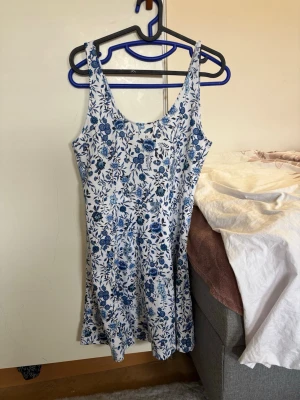 Blommig vit och blå sommarklänning - Söt kortklänning med blommigt mönster i blått och vitt. Klänningen är ärmlös med rundad halsringning och har en lätt utsvängd passform. Perfekt för varma dagar och har en fräsch, somrig look.
