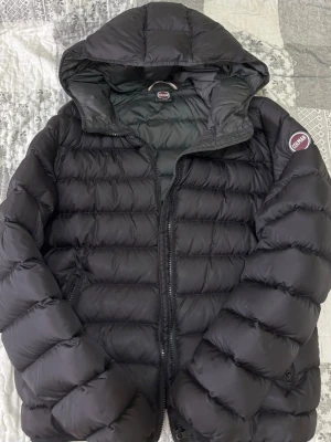 Svart pufferjacka från Colmar - Snygg svart pufferjacka från Colmar Originals med huva och dragkedja framtill. Jackan har en quiltad design och Colmar-logga på ärmen. Perfekt för kalla vinterdagar och riktigt skön att bära tack vare det vadderade materialet.