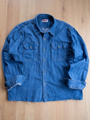 Blå jeansskjorta från Wrangler - Klassisk blå jeansskjorta från Wrangler med två bröstfickor och knappar framtill. Skjortan har lång ärm och är tillverkad i bomull. Perfekt för en avslappnad och cool stil.