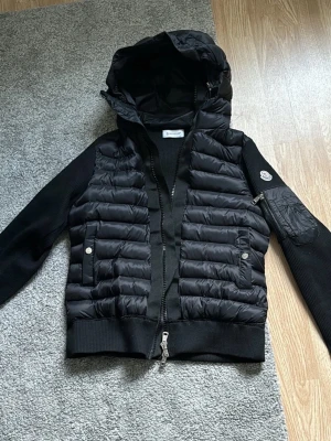 Svart dunväst med huva från Moncler - Snygg svart dunväst från Moncler med stickade ärmar och avtagbar huva. Västen har två fickor med tryckknappar och en Moncler-logga på ärmen. Perfekt för lager-på-lager och streetstyle. Materialet är en mix av dun och stickat tyg.