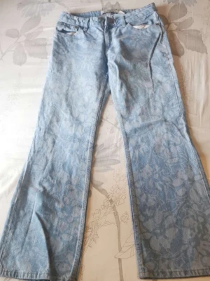 Ljusblå bootcut jeans med blommönster - Unika ljusblå jeans med diskret blommönster över hela tyget. Modellen har bootcut-ben och klassisk femficksdesign. Perfekta för dig som vill sticka ut med en cool och trendig look. Tillverkade i mjuk denim för skön känsla.