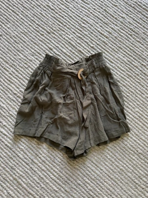 mörkgröna shorts med knyt och resår - Snygga beiga shorts med hög midja, resår i midjan och ett dekorativt knytband med trädetalj framtill. Luftig modell i mjukt linneliknande material, perfekta för varma dagar. Passar till både t-shirt och linne.