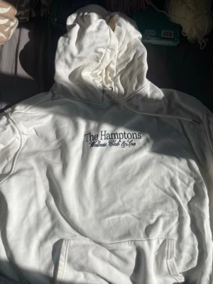Vit hoodie The Hamptons H&M - Säljer en vit hoodie från H&M med trycket 'The Hamptons Wellness Club & Spa' på bröstet. Hoodien har huva med dragsko och en stor magficka. Tillverkad i mjukt bomullsmaterial, perfekt för en avslappnad och stilren look.