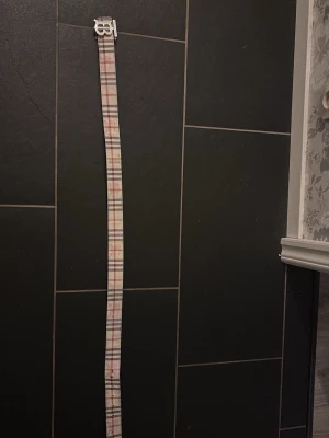 Burberry bälte med B-spänne - Snyggt bälte från Burberry med klassiskt rutmönster i beige, svart och rött. Bältet har ett silverfärgat B-spänne som ger en exklusiv känsla. Perfekt accessoar för att lyfta din outfit med en tidlös design.