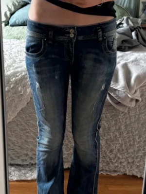 Blå bootcut jeans med slitningar - Säljer ett par blå bootcut jeans med snygga slitningar och dubbla knappar i midjan. Jeansen har klassiska fem fickor och ljusare tvättade partier framtill för en cool look. Perfekta för dig som gillar en avslappnad men trendig stil.
