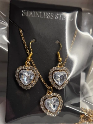 Guldigt smyckeset med hjärtan - Snyggt smyckeset i rostfritt stål med guldfärgade detaljer. Setet består av ett halsband och örhängen med hjärtformade hängen, dekorerade med gnistrande klara stenar och små kristaller runt om. Perfekt för dig som gillar bling och romantisk stil. Kedja 45 cm + förlängningskedja. Hänge ca 1,8 cm.