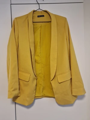 Senapsgul kavaj  - Snygg senapsgul kavaj med klassisk krage och två fickor framtill. Kavajen har en rak passform och är helfodrad. Perfekt för dig som vill addera färg till din outfit.