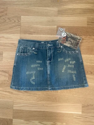 Lågmidjad kort jeanskjol  - Snygg lågmidjade jeans kjol med prislapp kvar. Cool design på bak och framsidan. Storlek 28. Från märket Rude Jeans. Midjemått: 39cm Längd fram: 32cm  Längd bak: 33cm 