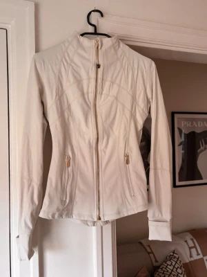 Vit vindjacka med dragkedja - Snygg vit vindjacka med figurnära passform och hög krage. Jackan har hel dragkedja framtill och två fickor med dragkedja. Dekorativa sömmar ger en sportig look. Passar S och M 