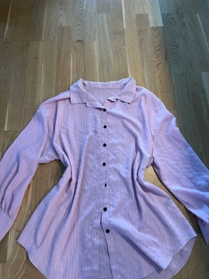 Ljusrosa skjorta från SHEIN - Säljer en ljusrosa skjorta från SHEIN i storlek XXL. Skjortan har långa ärmar, klassisk krage och svarta knappar framtill. Materialet är lätt och luftigt, perfekt för en avslappnad look. Passformen är loose och skjortan har en diskret randig struktur.