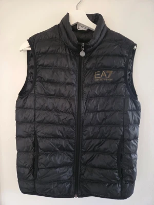 Svart dunväst från EA7 Emporio Armani - Snygg svart dunväst från EA7 Emporio Armani med guldfärgad logga på bröstet. Västen har hög krage, dragkedja framtill och två sidofickor. Tillverkad i glansigt material med quiltad design som ger en sportig och modern look.