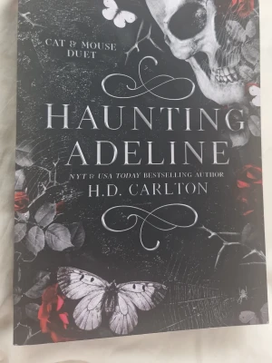 Haunting Adeline av H.D. Carlton - En mörk och mystisk pocketbok med svart omslag, dekorerad med vita fjärilar, röda rosor och en dödskalle. Boken är första delen i Cat & Mouse Duet och blandar skräck, romantik och spänning. Perfekt för dig som gillar psykologiska thrillers och gotisk estetik.