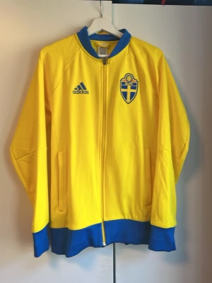 Gul Sverige adidas träningsjacka - Säljer en gul träningsjacka från adidas med blå detaljer och Sveriges landslagsemblem på bröstet. Jackan har hel dragkedja, två fickor och blå ribbade muddar. På ryggen står det 'SVERIGE' i blått. Perfekt för dig som vill stötta landslaget.