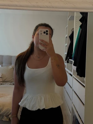 Vit volang topp med breda axelband - Säljer en söt vit topp med breda axelband och volang nedtill. Toppen har en figurnära passform och ärmlös design, perfekt för varma dagar. Tillverkad i mjuk bomull som känns skön mot huden. Enkel men ändå stylish detalj till din garderob. 