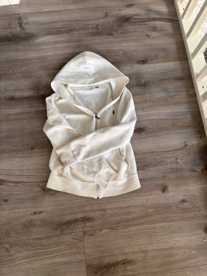 Vit hoodie från Polo Ralph Lauren - Säljer en vit hoodie från Polo Ralph Lauren med dragkedja framtill och klassisk huva. Hoodien har fickor på magen och den ikoniska lilla loggan broderad på bröstet. Tillverkad i mjuk bomull som är skön att ha på sig. Perfekt för en clean och stilren look.