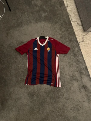 Randig Djurgården matchtröja Adidas - Säljer en Djurgården matchtröja från Adidas med breda marinblå och vinröda ränder, vitt krage och vita detaljer på sidorna. Tröjan har klubbmärke på bröstet och är kortärmad, perfekt för fotboll eller träning.
