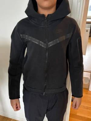 Svart Nike hoodie med dragkedja - Säljer en svart hoodie från Nike med hel dragkedja framtill och huva. Hoodien har ett diskret Nike-logga på bröstet och en snygg V-formad detalj i grått över bröstet. Ficka med dragkedja på ena ärmen och ribbade muddar. Perfekt för en sportig och avslappnad stil.