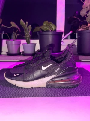 Nike Air Max 270 svarta sneakers - Säljer ett par svarta Nike Air Max 270 sneakers med vit sula och synlig Air-enhet i hälen. Skorna har mesh-överdel, vita detaljer och logga på sidan samt dragflik bak. Perfekta för dig som gillar sportig och modern stil.