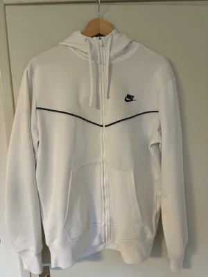Vit zip hoodie från Nike - Snygg vit hoodie från Nike med dragkedja framtill och svart Nike-logga på bröstet. Hoodien har en huva med dragsko, två stora fickor fram och en tunn svart linje som detalj över bröstet. Perfekt för en avslappnad och sportig stil.