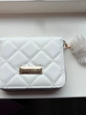 Vit quiltad plånbok med fluffig detalj - Snygg vit plånbok i quiltat mönster med guldfärgad dragkedja och en fluffig nyckelring. Insidan är ljusrosa med flera fack för kort och myntfack med dragkedja. Perfekt storlek för att ha med sig överallt och ger en lyxig känsla.