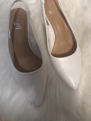 Vita pumps med öppen häl från VOX - Stilrena vita pumps från VOX med spetsig tå och öppen häl. Skorna har en smal klack och är tillverkade i veganskt material. Perfekta för dig som vill ha en elegant och modern look.