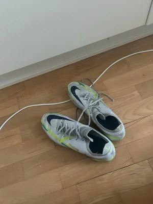 Nike Phantom vita fotbollsskor - Säljer ett par Nike Phantom fotbollsskor i vitt med neon- och mörkblå detaljer. Skorna har snörning och dobbar för spel på gräs. Ovandelen är i ett lätt syntetmaterial som ger bra bollkontroll och passform. Perfekta för dig som vill ha snabba och snygga skor på planen.
