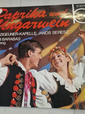 Paprika und Ungarwein LP - LP-skiva med titeln 'Paprika und Ungarwein' av Zigeuner-Kapelle Janos Seresz och Sari Barabas. Omslaget har färgstark design med folkdräkter och tydlig text i rött och vitt. Skivan innehåller traditionell ungersk musik och sång. Material: vinyl. Perfekt för dig som gillar klassisk och folkmusik.