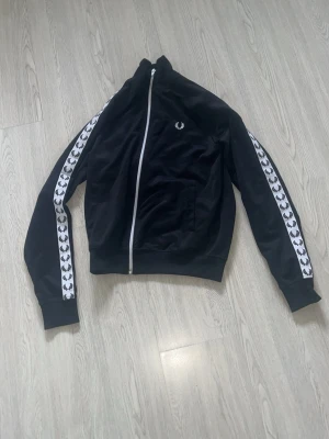 Svart track jacket från Fred Perry - Säljer en svart track jacket från Fred Perry med vit dragkedja och klassiska loggan broderad på bröstet. Ärmarna har vita band med Fred Perry-kransar längs hela sidan. Perfekt för en sportig och stilren look.